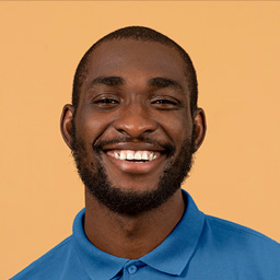 Portrait of a smiling man with a beard wearing a blue polo shirt against a light brown background.