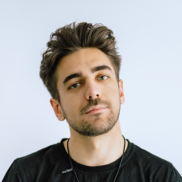 Young man with styled dark hair and light beard wearing a black shirt and earphones against a plain light background.