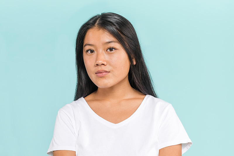 Portrait of a young woman with long black hair wearing a white t-shirt against a light blue background.