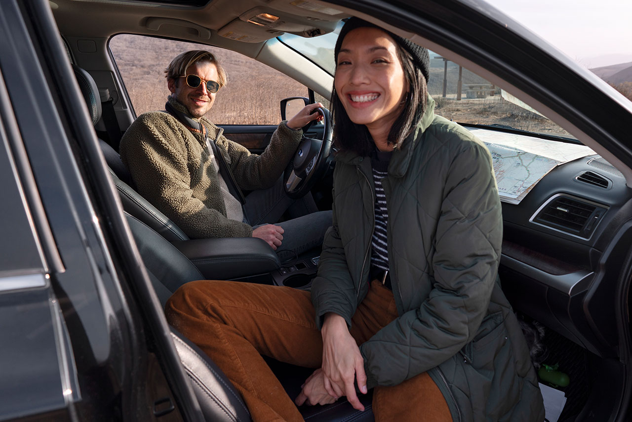 Smiling man and woman sitting inside a car with a map on the dashboard, dressed in warm outdoor clothing.