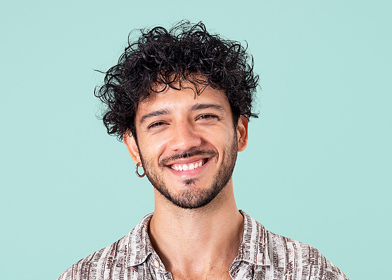 Smiling man with curly dark hair and a beard wearing a patterned shirt against a light blue background.
